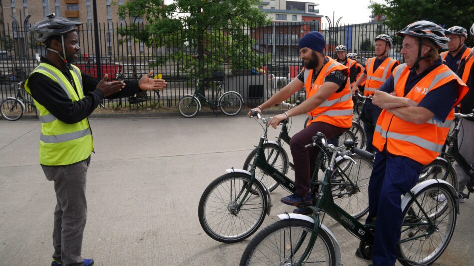 Group of freight drivers in orange hi-vis and on cycles receiving instruction from a cycle instructor