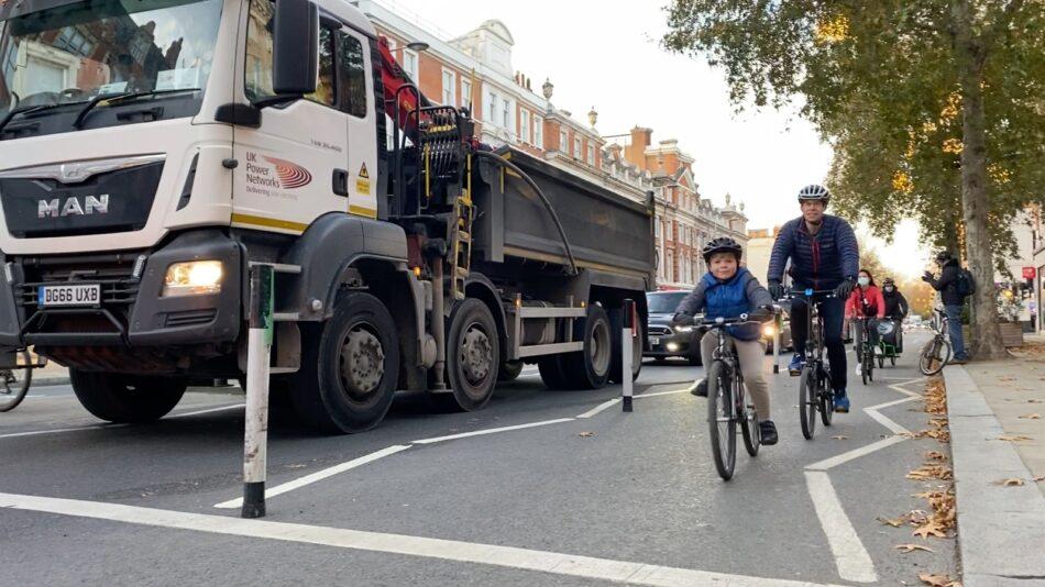 A small child on a bike on Kensington High Street, on a protected bike lane, with bollards between him and a lorry in the lane alongside