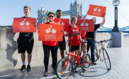 A group of LCC activists smiling and holding up red LCC signs. Tower Bridge is visible behind them.