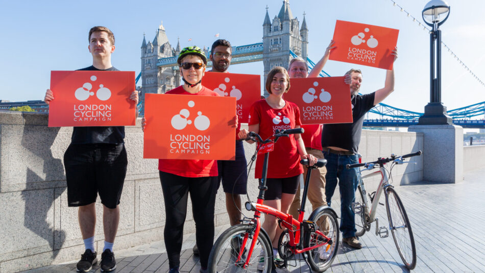 A group of LCC activists smiling and holding up red LCC signs. Tower Bridge is visible behind them.