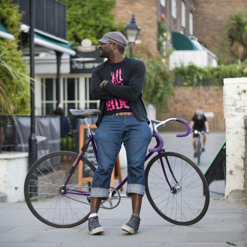 A man in casual clothing and a cap leaning against a fixie bike and smiling