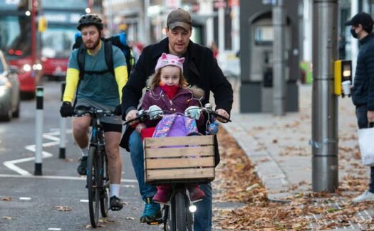 A man cycling with a young girl in a front seat, and her purple backpack in the front basket