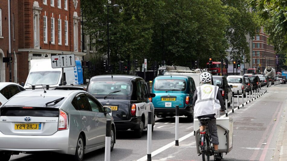 Euston Road cycle track made with semi-separated 'wand' protection, but image now flipped horizontally, so on the left, cars and taxis queue in congested traffic, to the right a cargo bike delivery cyclist rides away from camera.