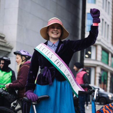 Woman raising fist and holding bike in front of her dressed as a suffragette at LCC protest ride