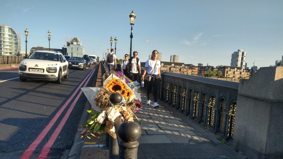 Flowers in foreground tied to railing of Battersea Bridge, motor traffic and bridge in background, following fatal collision with 27 year old woman cycling on 10 August 2023